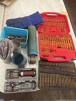 View of assorted drill bits in red and black cases, blue welding gloves, welding electrodes container, and small metal tools on table