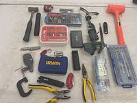 Overall view of assorted tools spread on a white surface, showing all items in the lot including hammers, pliers, drill, and small containers with screws and drill bits.