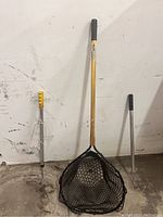 Photo of fishing net with black mesh and gold handle, with two fishing gaffs (yellow and black grips) against a white wall.
