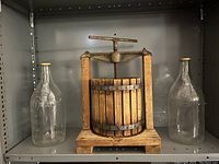 Front view on shelf of antique wooden fruit press juicer flanked by two large glass bottles with screw lids.
