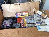 Box with curved HO scale track pieces, small model building with red roof, plastic bags with scenery and miniature figures, and boxed powered locomotives.