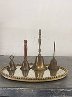 Four brass bells of different sizes and handle styles arranged on an oval mirrored plate with a gold-tone decorative edge.