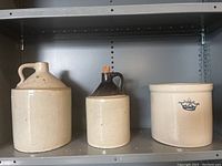 Photo showing three antique jugs on a metal shelf: a large beige jug with handle, a medium jug with dark brown top and cork stopper, and a small cylindrical crock with blue crown emblem.