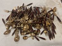 Overview of the large pile of assorted flatware with brown handles and brass colored metal parts on a white surface.