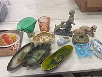 Photo showing an assortment of decorative home items including glass pitchers, embossed trays, ceramic bowl, porcelain clog, floral trinket dish, and porcelain figurines.