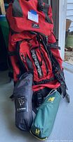 Red Neptune backpack with black and gray straps and buckles, positioned outdoors on a porch with the Helinox and Alite camping chair bags in front.
