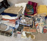 Photo showing assorted fabric pieces in various colors and patterns, various sewing tools including buttonhole cutter, belt buckles, ribbons, pins, and snaps arranged on floor.