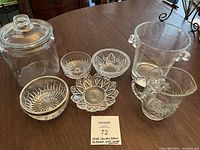 Glass jar with lid, small pitcher, ice bucket, and four small candy dishes arranged on wooden table