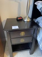 Photo showing the full black wooden nightstand with two drawers and an open shelf at bottom, positioned beside a bed. The drawers have round brass pulls. The surface holds a lamp and clock (not included).