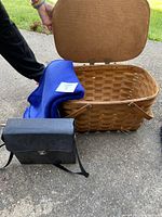 Photo shows open woven picnic basket with hinged lid and two handles, blue fleece blanket draped over basket edge, and black carrying case resting in front with shoulder strap visible.