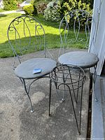 Two rod iron chairs with curved backs and gray seat cushions next to a round rod iron plant stand with grid top, placed outside on concrete surface.