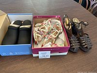 Three pairs of women's shoes on table including Dansko clogs in box, floral sandals in pink box, and brown wedge sandals
