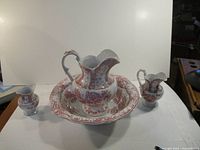 Full view of 4 piece wash bowl set, showing large bowl, large pitcher, smaller pitcher, and small vase-like piece, all with red floral pattern on white ceramic.