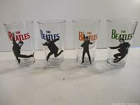Front view of four clear pint glasses each decorated with a colorful 'The Beatles' logo and black silhouettes of band members in various poses.