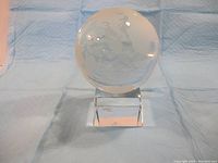 Glass globe on rectangular glass base stand photographed against a blue fabric backdrop. The globe is clear with etched world map details.