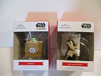 Two Hallmark Star Wars Christmas ornaments displayed in their packaging. Left is Grogu in his hover pram. Right is Obi-Wan Kenobi in Jedi robes with a blue lightsaber.