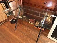 Side view of rectangular glass top table with chrome crossbar base on hardwood flooring.