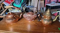 Three copper kettles displayed on a wooden surface with various background items, showing their relative sizes and handle styles.