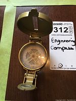 Top view of the open engineering compass showing the compass face, needle, cardinal points, and hinged lid.