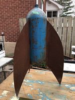 View of the metal farm yard art shaped like a rocket, showing blue cylindrical body and rusted large metal fins, sitting on a weathered surface outdoors.