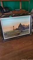 Painting with barn and windmill landscape placed on wooden floor, showing full frontal view and ornate frame.