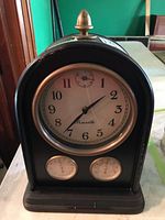 Front view of Penrith mantel clock showing white dial with black Arabic numerals, smaller second dial, and two additional meters below. Brass finial on top of wooden case visible.