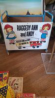 Front and side view of the Raggedy Ann and Andy wooden toy cart showcasing the colorful illustrations and metal frame.