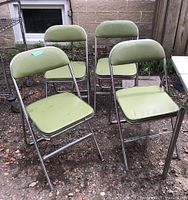 Four retro folding chairs with light green vinyl seats and chrome frames shown outdoors on ground with some dirt and stones.