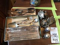 Assortment of silver-plated cutlery including serving spoons, child’s cutlery, salt and pepper shakers and napkin rings displayed on a wooden table.