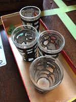 Four small clear glasses with frosted finish and detailed sterling silver overlay featuring religious and abstract geometric designs, displayed top-down and inside box.