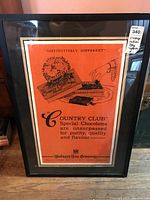 Framed vintage advertising tapestry in black frame with orange background and text for Hudson's Bay Country Club Special Chocolates.