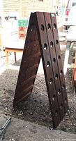 Full view of the dark stained wooden A-frame wine rack standing upright outdoors, showing two wooden panels with bottle neck holes and a chain for support.
