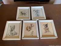 Five vintage puppy dog paintings arranged on wooden surface, each in white wood frame.