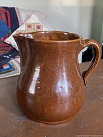 Front and side view of the brown glazed earthenware pitcher jug placed on a wooden table with fabric couch background.