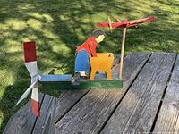 Side angle view of the antique wooden whirligig showing the washing lady figure, yellow washtub, red clothes drying rack, and front spinning windmill blades on a green base placed on an outdoor wooden table.