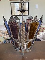 View of antique hanging light showing laurel leaf cast metal frame and stained glass panels in amber, blue, and ruby colors. One amber panel is missing.