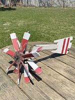 Side view of the antique wooden airplane whirligig on a wooden table outdoors, showing front double propellers and tail section with red stripes.