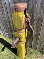 Front angle view showing wooden yellow pump column with attached rusted metal handle and spout, outdoor setting with wooden fence background.