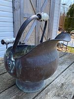 Front angled view of the hammered brass scuttle bucket showing the curved body, pedestal base, arched handle with Delft porcelain handle grips, and a sculpted lion head on the side.