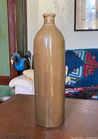 Full view of tall slender earthenware soda bottle standing on wooden surface with background showing part of a room.