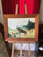 Full view of folk art elk painting with wooded lodge-style frame on wooden easel in front of red curtains.