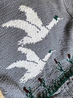 Close-up photo showing part of the knitted sweater with two flying white duck silhouettes and green cattail plants on a grey wool background.