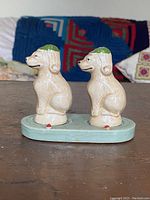 Side view of two cream-colored porcelain dog salt and pepper shakers with green hats on a pale green base with red dot detail