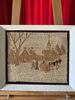Front view of hooked rug tapestry framed art showing a winter rural Quebec village scene with horses, houses and church steeple