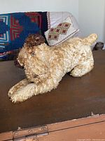 Side view of the vintage brown mohair stuffed dog laying down on a wooden surface. Background shows some textile items and art on the wall.