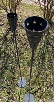 Two tall vintage black cast iron outdoor candle holders placed on grass, showing conical tops with triangular cutouts and circular flat base.