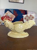 Side profile of yellow ceramic rooster planter showing shape, color, and size compared with couch.