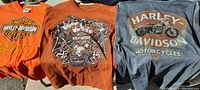 Three Harley Davidson T-shirts with various motorcycle and skull graphics in orange and gray colors