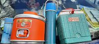 Photo showing four vintage insulated items: orange and white cooler, greenish blue cooler with red lid, tall blue thermos, smaller blue thermos on a reflective surface.
