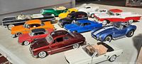 Top view of multiple diecast cars including blue Cobra convertible, Cadillac with broken pieces, various classic models in multiple colors.
